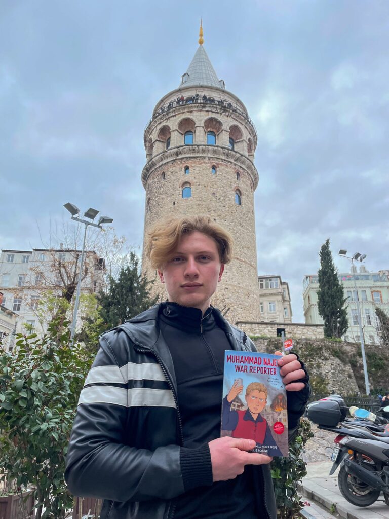 Muhammad Najem stands in a Syrian city holding a copy of the graphic novel Muhammad Najem, War Reporter.
